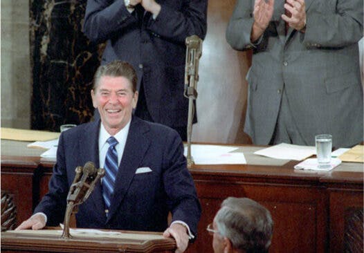 Ronald Reagan stands behind a podium. George H. W. Bush and Tip O'Neill stand behind him and clap. An American flag hangs behind them.